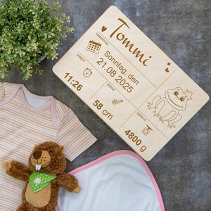 以下が含まれることがあります： 「Tommi」と刻まれた木製のプレート。プレートには、日付「21.08.2025」、時刻「11:26」、体重「4800 g」、身長「58 cm」も刻まれています。