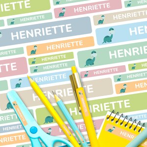 May include: A set of colourful personalised name labels featuring a cartoon dinosaur design. The labels are printed with the name "Henriette" in white text. The labels are ideal for school supplies, lunchboxes, and other items.
