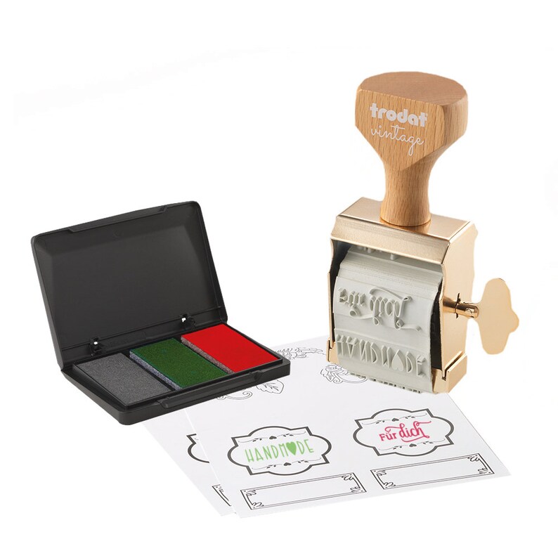 K&ouml;nnte beinhalten: Ein goldfarbener Vintage-Stempel mit Holzgriff und dem Text "trodat vintage". Der Stempel hat ein Design mit einem Herzen mit dem Text "Handmade" und einem Herzen mit dem Text "F&uuml;r dich". Der Stempel wird mit einem schwarzen Stempelkissen mit drei Farben geliefert: Grau, Gr&uuml;n und Rot. Der Stempel wird au&szlig;erdem mit mehreren wei&szlig;en Etiketten geliefert, auf denen der Text "Handmade" und "F&uuml;r dich" gedruckt ist.