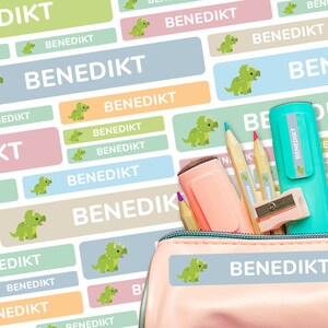 May include: A pink pencil case with a blue and white label that says "BENEDIKT". The label has a green dinosaur illustration. The pencil case is open and has pencils, a sharpener, and a highlighter inside.