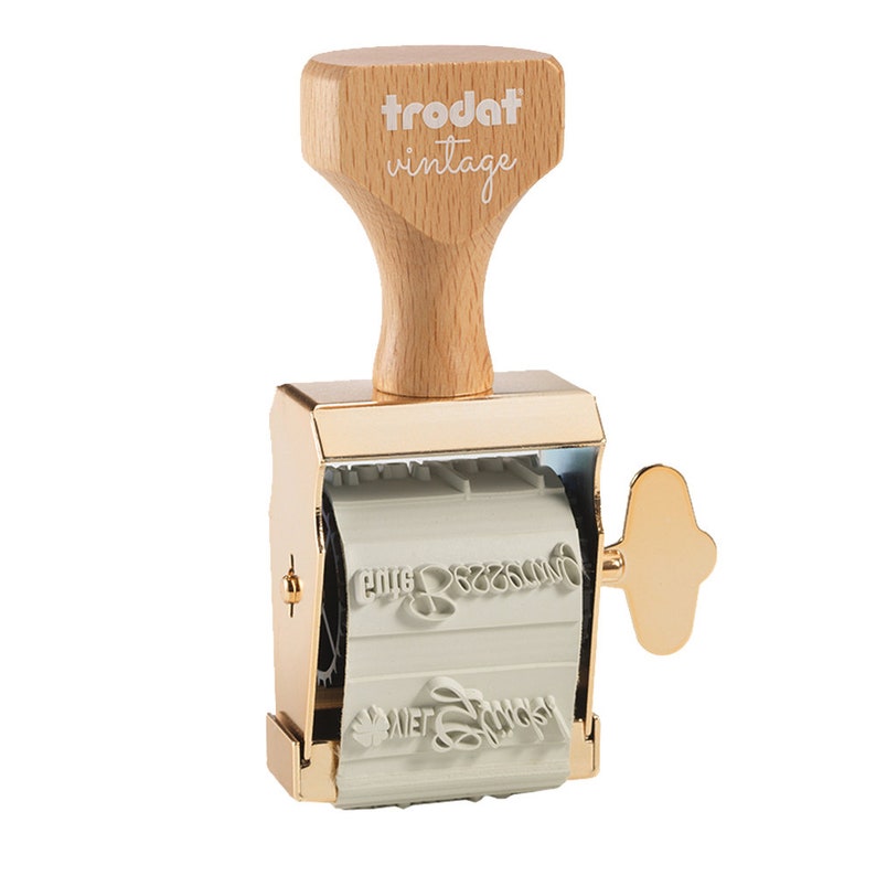 Puede incluir: Un sello vintage Trodat de color dorado con un mango de madera. El sello tiene un mecanismo giratorio con varias l&iacute;neas de texto.