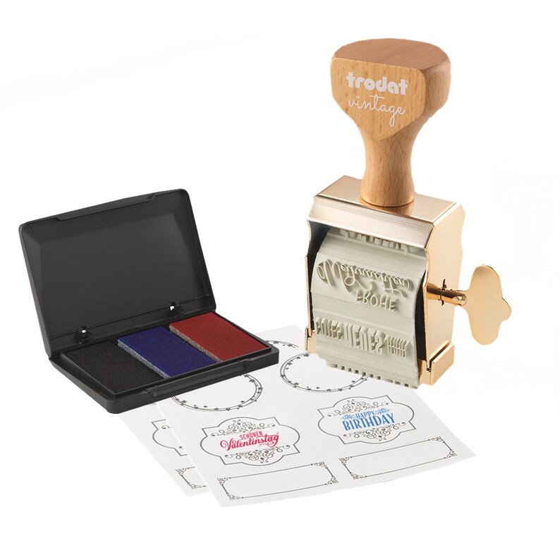 Puede incluir: Un sello de goma vintage de color dorado con mango de madera, una almohadilla de tinta negra con tres colores de tinta y varias etiquetas blancas en blanco con dise&ntilde;os preimpresos. El sello presenta el texto "trodat vintage" y el texto "Made in Germany".