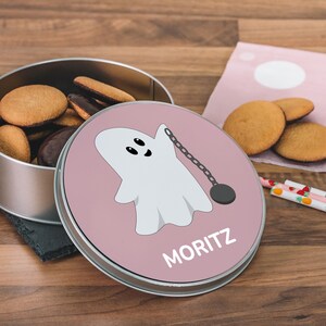 Puede incluir: Una lata redonda de galletas de metal con una tapa rosa que presenta un fantasma de dibujos animados sosteniendo un peso en una cadena. La tapa tiene el nombre "MORITZ" impreso. La lata está llena de galletas.