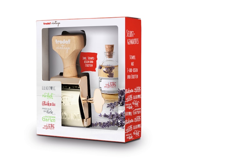K&ouml;nnte beinhalten: Ein goldfarbener Stempel mit einem Holzgriff und eine klare Glasflasche mit einem Korkverschluss. Der Stempel hat ein wei&szlig;es Etikett mit dem Text "HANDMADE" und "LIEBE" in Rot. Die Flasche hat ein wei&szlig;es Etikett mit dem Text "LIEBE" und "gemacht". Der Hintergrund ist wei&szlig;.
