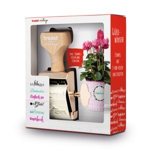May include: A gold-colored self-inking stamp set with a wooden handle and a white ink pad. The stamp set comes with three different messages: "Ich lieb dich", "Dankeschön", "Einfach so". The set also includes a small potted plant with pink flowers and a white pot with a pink and white pattern.