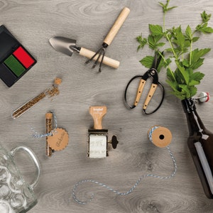 K&ouml;nnte beinhalten: Ein Holzstempel mit einem Metallrahmen und einem wei&szlig;en Etikett mit der Aufschrift "trodat vintage". Der Stempel befindet sich auf einer Holzoberfl&auml;che mit anderen Bastelutensilien, darunter ein kleines Glasfl&auml;schchen, eine Schere, eine kleine Schaufel, eine Garnrolle und eine Bierflasche.