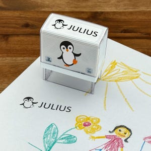 Könnte beinhalten: Ein weißer Stempel mit Selbsttinte, einem Pinguin-Motiv und dem Namen "JULIUS" darauf gedruckt. Der Stempel befindet sich auf einem weißen Blatt Papier mit einer Kinderzeichnung von einer Sonne, einer Blume und einer Person.