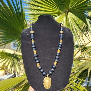 May include: A necklace featuring dark metallic beads interspersed with patterned yellow and orange cylindrical beads, accented by small gold-toned spacers. The centerpiece is a gold-toned pendant shaped like a stylized human face with textured hair.