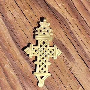 May include: A gold-toned metal pendant in the shape of an ornate cross featuring intricate geometric lattice patterns, a small loop at the top for suspension, and symmetrical decorative extensions on the horizontal and vertical arms of the cross.