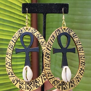 May include: A pair of oval-shaped wooden earrings featuring a black ankh symbol centered within each frame. The outer edges of the wood are painted gold with black geometric patterns. A single white cowrie shell is attached to the base of each ankh.