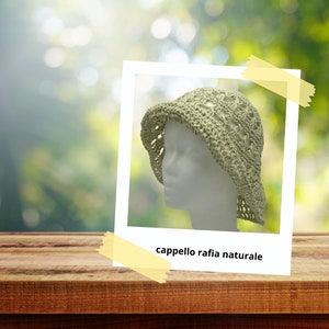 Pode incluir: Chapéu de crochê verde claro com aba, exibido em uma cabeça de manequim branca. O chapéu está sobre um fundo desfocado de folhagem verde e uma superfície de madeira. O texto "cappello rafia naturale" é visível.