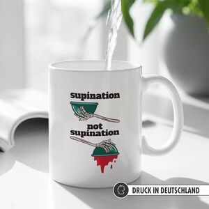 May include: White ceramic mug with a black and green illustration of a skeleton hand holding a bowl. The text "supination" and "not supination" is printed on the mug. The mug has a black handle and is filled with water.