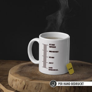 May include: White ceramic mug with a black scale printed on the side. The scale has text in German that reads "Bitte nicht ansprechen! Immer noch nicht! Fast wach Ja bitte? Zu sp&auml;t! Ich muss aufs Klo..."  The mug is on a wooden surface with a tea bag.