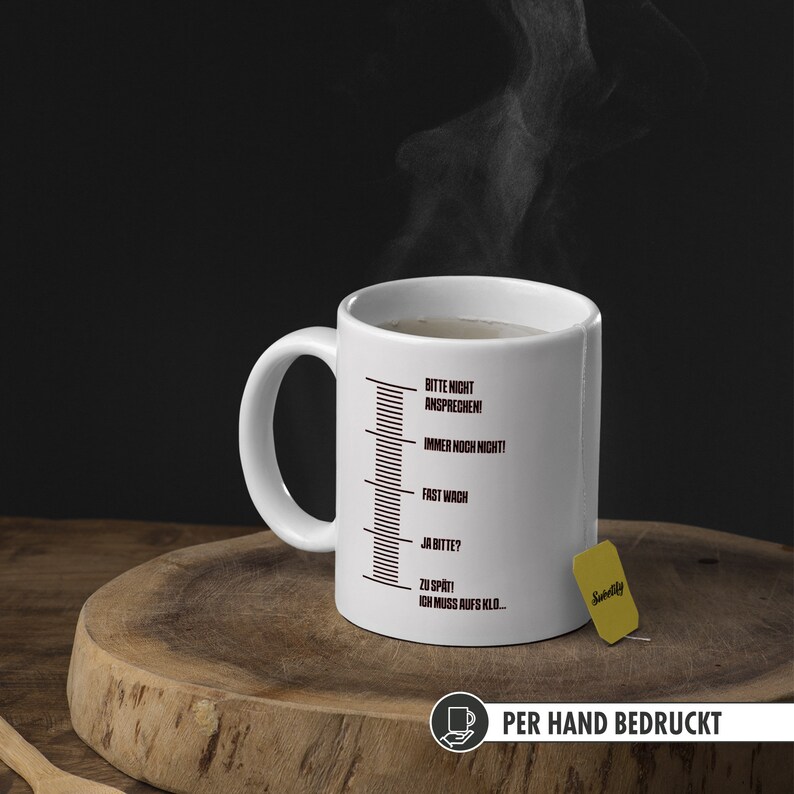 May include: White ceramic mug with a black scale printed on the side. The scale has text in German that reads "Bitte nicht ansprechen! Immer noch nicht! Fast wach Ja bitte? Zu sp&auml;t! Ich muss aufs Klo..."  The mug is on a wooden surface with a tea bag.