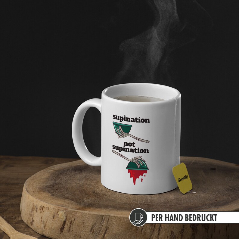 May include: White ceramic mug with a black and green illustration of a skeleton hand holding a bowl. The text "supination" and "not supination" is printed on the mug. The mug is filled with a brown liquid and a tea bag is on the table next to the mug.