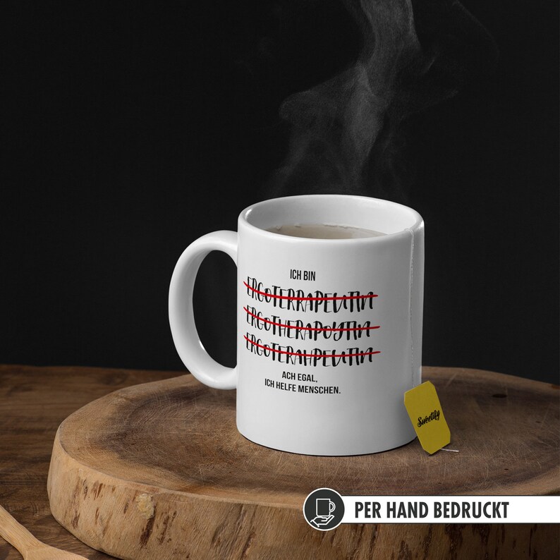 May include: White ceramic mug with black text that reads "ICH BIN" followed by three lines of text with the word "ERGOTERAPELUTIA" crossed out. The last line reads "ACH EGAL. ICH HELFE MENSCHEN."  The mug is sitting on a wooden surface with a yellow tea bag.