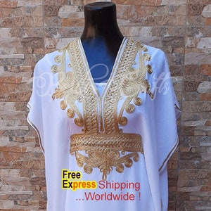 May include: White kaftan with gold embroidery. The embroidery features a geometric pattern with a central design that resembles a stylized flower. The kaftan has a V-neckline and short sleeves.