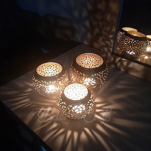 May include: Three gold metal candle holders with intricate cut-out designs. The candle holders are lit and casting shadows on the surface they are on.