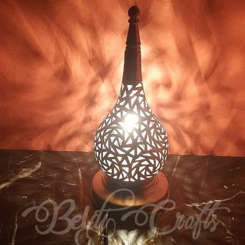 Puede incluir: Una l&aacute;mpara de mesa decorativa con una base de metal y una pantalla con un patr&oacute;n geom&eacute;trico. La l&aacute;mpara est&aacute; encendida y proyecta un brillo c&aacute;lido.