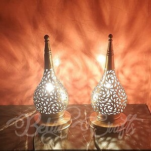 May include: Two decorative metal lamps with intricate cut-out designs. The lamps are gold in color and have a pear-shaped base. They are lit and casting a warm glow.