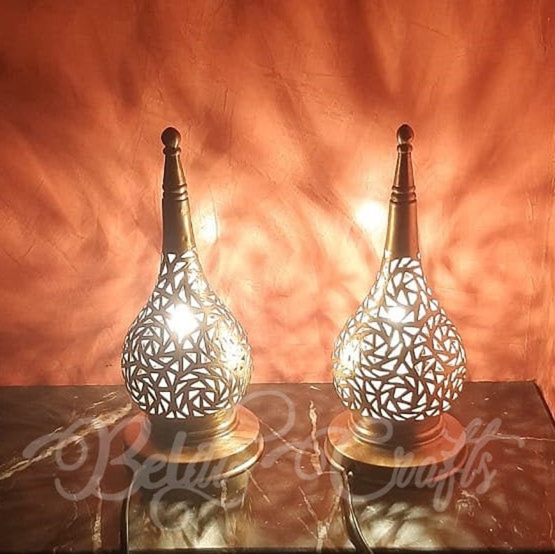 May include: Two decorative metal lamps with intricate cut-out designs. The lamps are gold in color and have a pear-shaped base. They are lit and casting a warm glow.