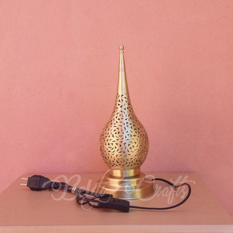 Puede incluir: Una l&aacute;mpara de mesa de metal dorado con un patr&oacute;n geom&eacute;trico decorativo, intrincado y delicado. La l&aacute;mpara tiene una base en forma de l&aacute;grima y una parte superior puntiaguda.