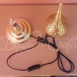 Puede incluir: Una l&aacute;mpara de mesa de metal dorado con una pantalla redonda perforada y un cable con un interruptor en l&iacute;nea. La l&aacute;mpara est&aacute; sobre una superficie de madera.