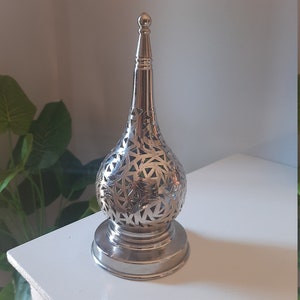 May include: A silver metal candle holder with a decorative, intricate, cut-out design. The holder has a round base and a tall, pointed top.