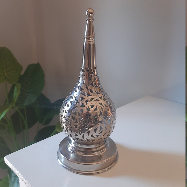 May include: A silver metal candle holder with a decorative, intricate, cut-out design. The holder has a round base and a tall, pointed top.