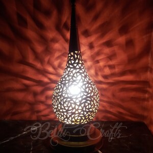 Puede incluir: Una l&aacute;mpara de mesa decorativa con un dise&ntilde;o geom&eacute;trico recortado en forma de l&aacute;grima. La l&aacute;mpara est&aacute; hecha de metal y tiene un acabado dorado c&aacute;lido. La luz brilla a trav&eacute;s de los recortes, creando un hermoso patr&oacute;n en la pared detr&aacute;s de ella.