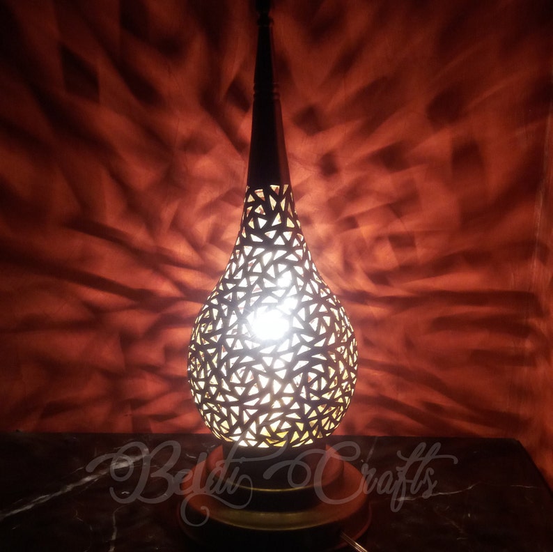 Puede incluir: Una l&aacute;mpara de mesa decorativa con un dise&ntilde;o geom&eacute;trico recortado en forma de l&aacute;grima. La l&aacute;mpara est&aacute; hecha de metal y tiene un acabado dorado c&aacute;lido. La luz brilla a trav&eacute;s de los recortes, creando un hermoso patr&oacute;n en la pared detr&aacute;s de ella.