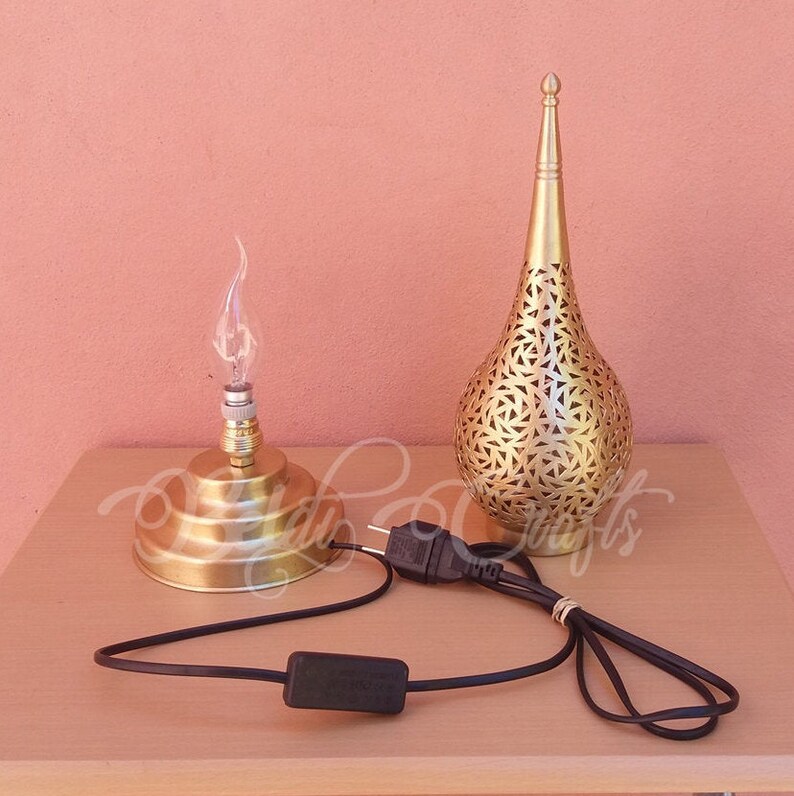 Puede incluir: Una l&aacute;mpara de mesa de metal dorado con un dise&ntilde;o decorativo perforado. La l&aacute;mpara tiene una base redonda y una pantalla alta y puntiaguda. La l&aacute;mpara est&aacute; enchufada y tiene un interruptor en el cable.