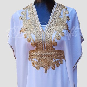 May include: White kaftan with gold embroidery. The embroidery features intricate designs around the neckline and waist.