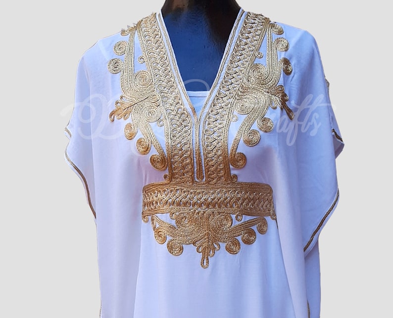 May include: White kaftan with gold embroidery. The embroidery features intricate designs around the neckline and waist.