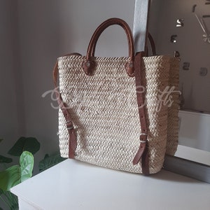 May include: A woven straw tote bag with brown leather straps and buckles. The bag has a rectangular shape and is made of a natural, light-coloured material.