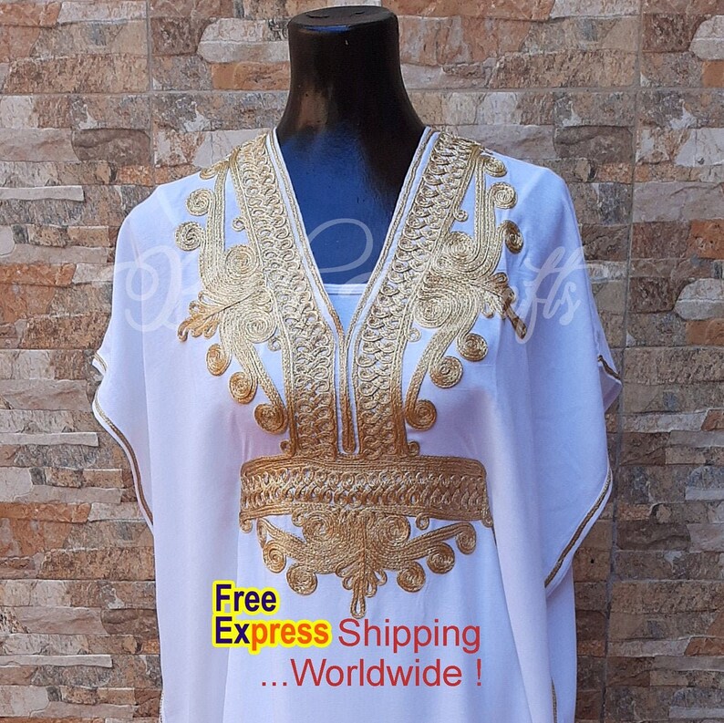 May include: White kaftan with gold embroidery. The embroidery features a geometric pattern with a central design that resembles a stylized flower. The kaftan has a V-neckline and short sleeves.