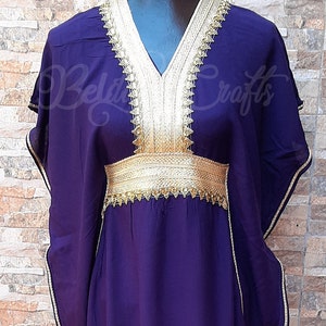May include: A long, purple kaftan with gold trim around the neckline, sleeves, and waist. The kaftan has a V-neckline and is made of a soft, flowing fabric.