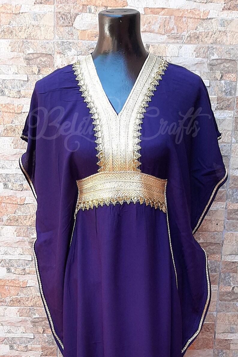 May include: A long, purple kaftan with gold trim around the neckline, sleeves, and waist. The kaftan has a V-neckline and is made of a soft, flowing fabric.