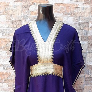 May include: A purple kaftan with gold embroidery around the neckline and waist. The kaftan has a V-neck and long sleeves.