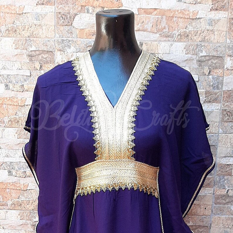 May include: A purple kaftan with gold embroidery around the neckline and waist. The kaftan has a V-neck and long sleeves.