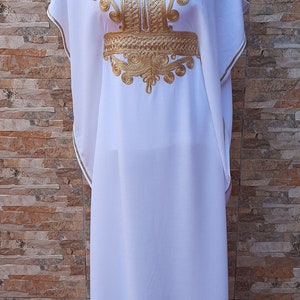 May include: A white kaftan with gold embroidery. The embroidery features intricate floral and geometric patterns. The kaftan has a V-neckline and short sleeves.