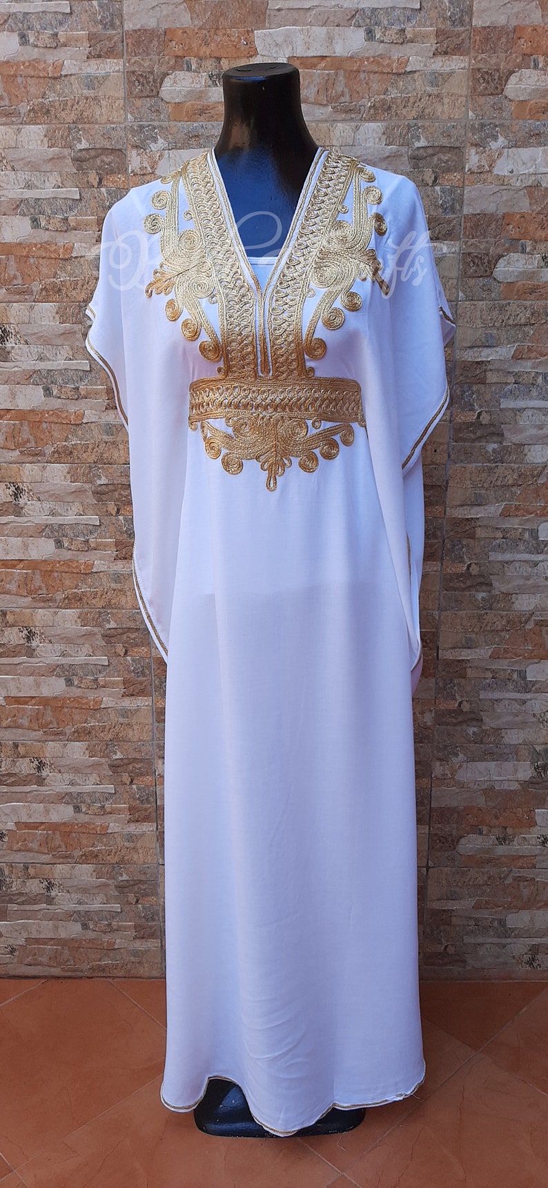 May include: A white kaftan with gold embroidery. The embroidery features intricate floral and geometric patterns. The kaftan has a V-neckline and short sleeves.