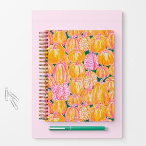 Pu&ograve; includere: Un taccuino a spirale con un motivo ripetuto di zucche gialle e arancioni con accenti rosa su sfondo verde scuro, posizionato su una base rettangolare rosa chiaro con tre graffette argentate e una penna verde.