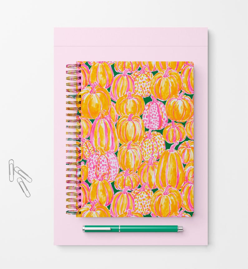 Pu&ograve; includere: Un taccuino a spirale con un motivo ripetuto di zucche gialle e arancioni con accenti rosa su sfondo verde scuro, posizionato su una base rettangolare rosa chiaro con tre graffette argentate e una penna verde.