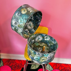 May include: A vintage hat box with a floral print in shades of blue, green, and white. The box is open and the lid is resting on a yellow chair. The box is sitting on a black and red floral rug.