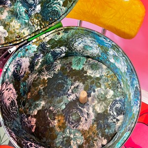 May include: A vintage floral patterned hat box with a green lining and a cork stopper in the center. The box is open and the inside is visible.