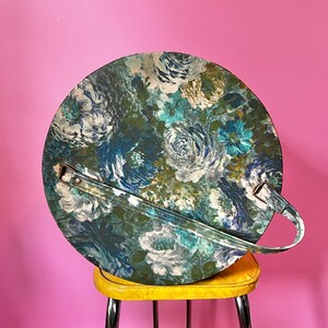 May include: A round, floral patterned storage container with a brown handle. The container is made of a hard, plastic-like material and has a blue, green, and white floral design.
