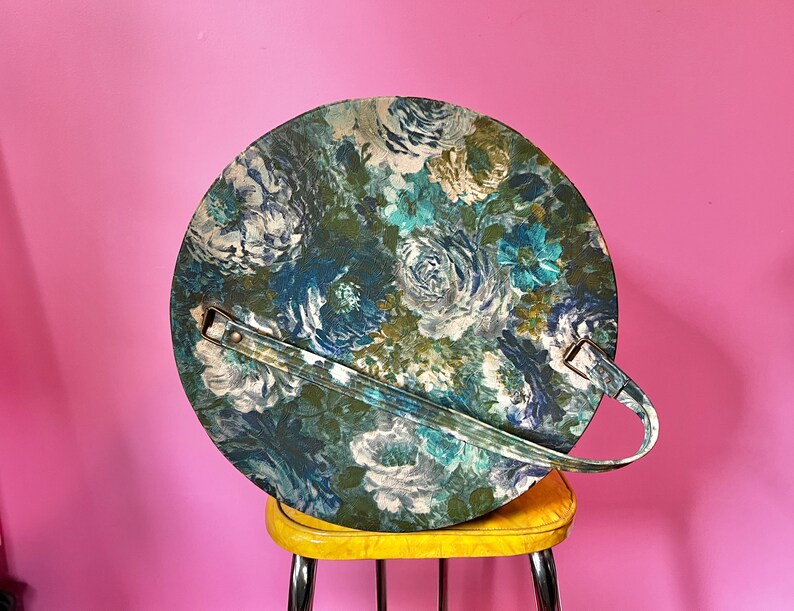 May include: A round, floral patterned storage container with a brown handle. The container is made of a hard, plastic-like material and has a blue, green, and white floral design.