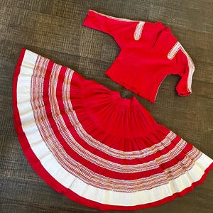 May include: A red skirt with white and gold trim and a matching red top with gold trim. The skirt is full and has multiple layers. The top has long sleeves and a round neckline.