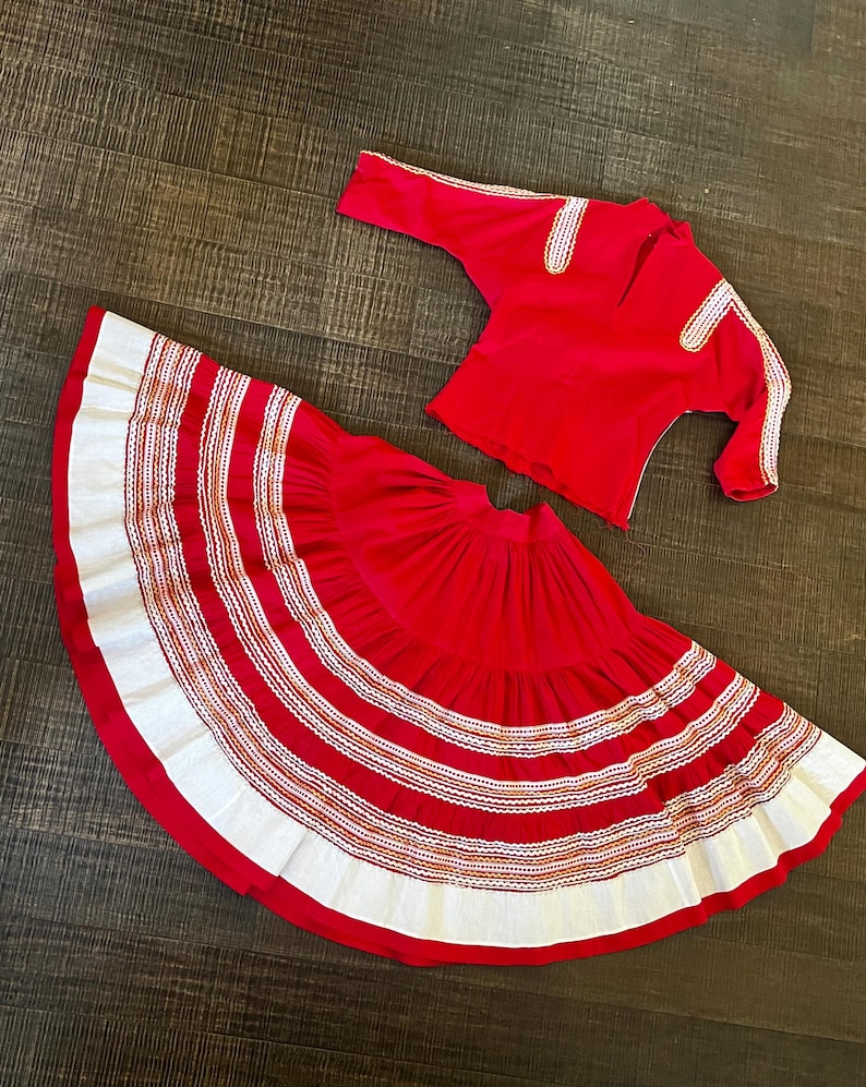 May include: A red skirt with white and gold trim and a matching red top with gold trim. The skirt is full and has multiple layers. The top has long sleeves and a round neckline.